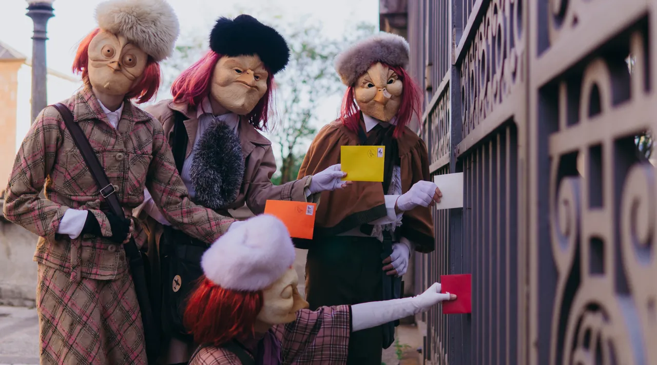 Figuras mascaradas entregam cartas coloridas junto a um portão de ferro