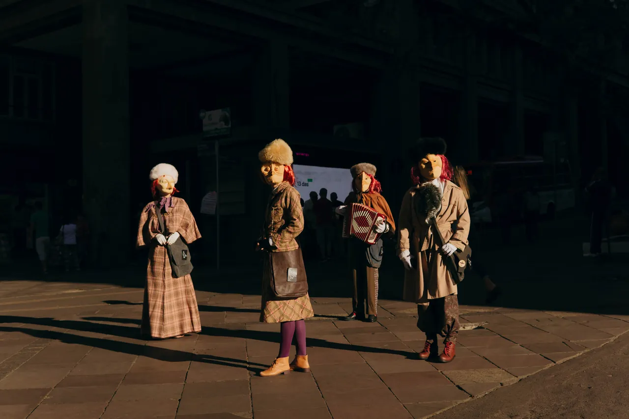 Quatro figuras mascaradas em pé numa praça escura, com sol rasante no chão