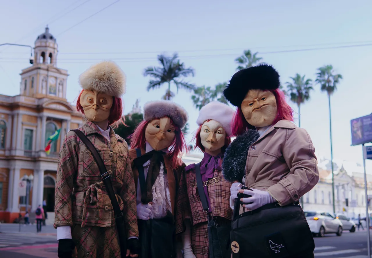 Quatro figuras mascaradas diante de um prédio histórico com palmeiras